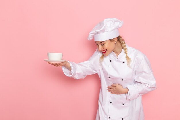 Front view young female cook in white cook suit holding cup of coffee with smile on the pink space cook  