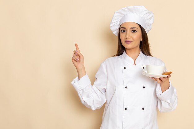 Front view of young female cook in white cook suit holding cup with coffee on white wall