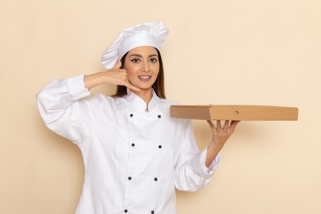Front view of young female cook in white cook suit holding food box on light-white wall