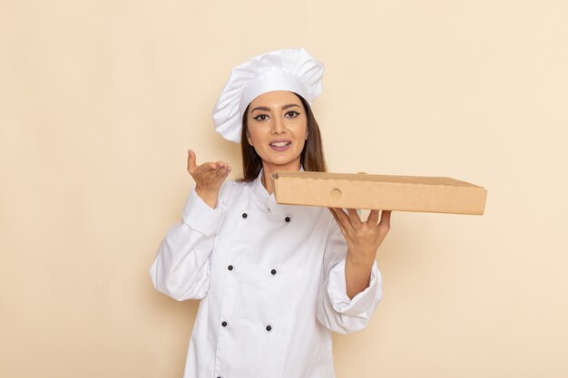 明るい白い壁にフードデリバリーボックスを保持している白いクックスーツの若い女性料理人の正面図