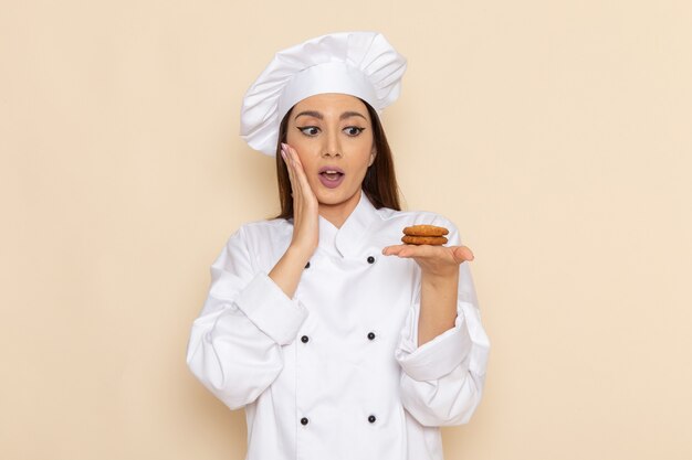 Front view of young female cook in white cook suit holding little cookies on the light-white wall