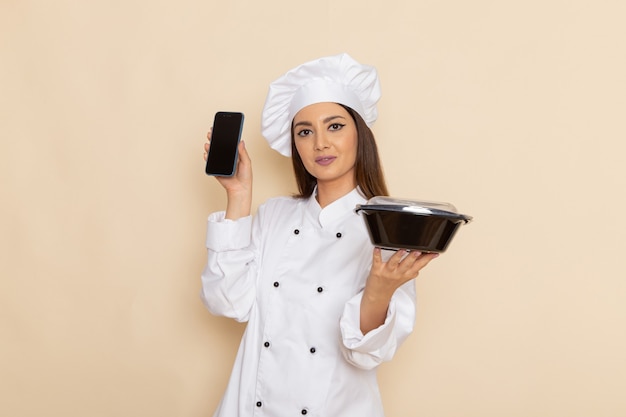 Vista frontale del giovane cuoco femminile in vestito bianco da cuoco che tiene telefono e ciotola nera sulla parete bianco-chiaro