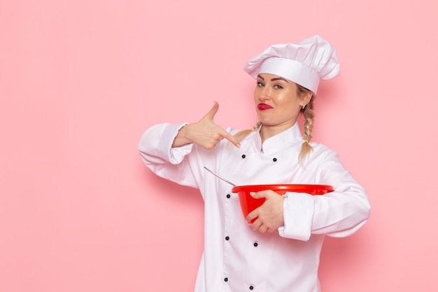 Front view young female cook in white cook suit holding red plastic bowl on the pink space cook  food meal