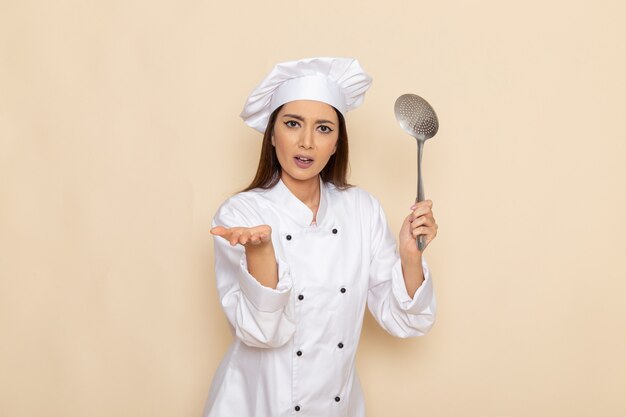 Front view of young female cook in white cook suit holding silver big spoon on light white wall