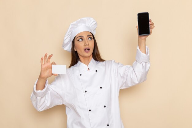 Front view of young female cook in white cook suit holding smartphone and card on light-white wall