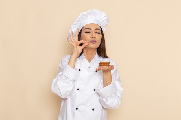 Front view of young female cook in white cook suit holding sweet cookies on light-white wall