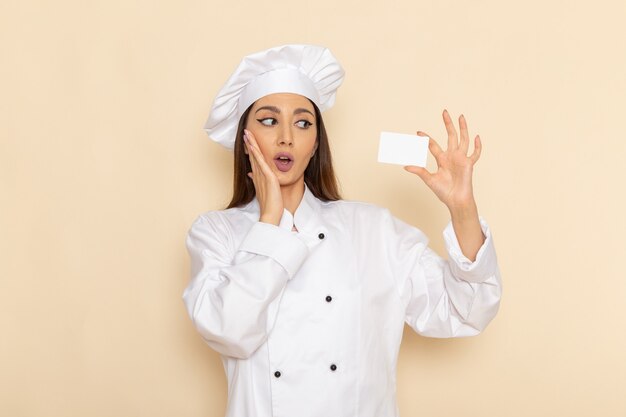 明るい白い机の上の白いカードを保持している白いクックスーツの若い女性料理人の正面図調理キッチン仕事仕事料理労働者