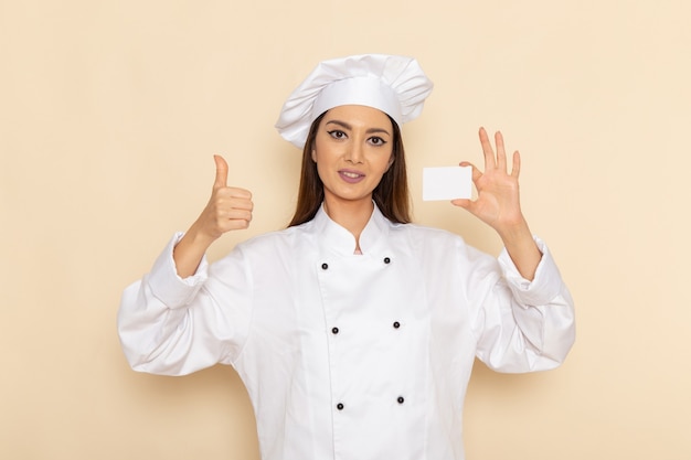 Vista frontale del giovane cuoco femminile in vestito bianco cuoco che tiene scheda bianca sulla parete bianco-chiaro