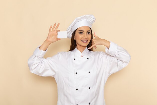 Front view of young female cook in white cook suit holding white card on light-white wall