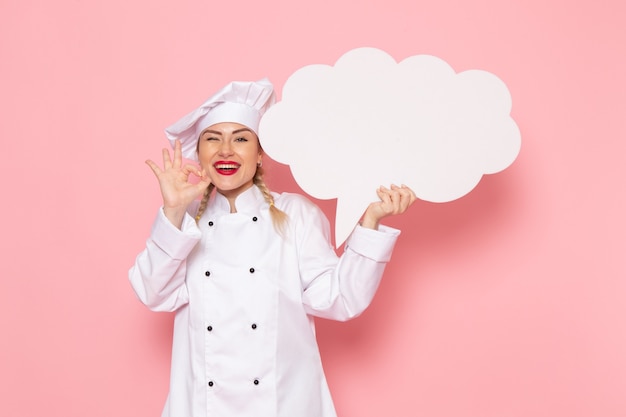 Free photo front view young female cook in white cook suit holding white sign with slight smile on the pink space cook   photo