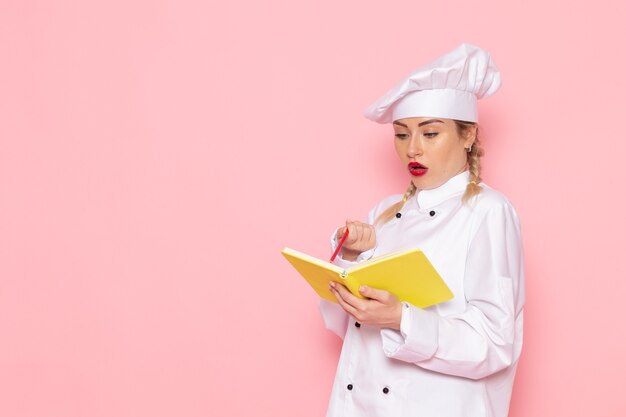 ピンクのスペースクック食品に黄色のコピーブックと鉛筆を保持している白いクックスーツで正面の若い女性クック
