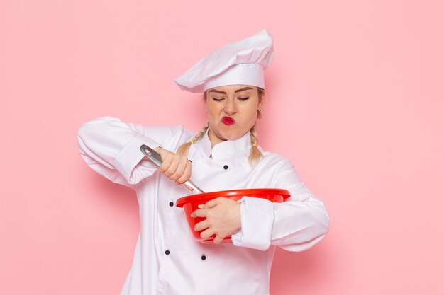 Вид спереди молодая женщина-повар в белом костюме повара, смешивающая красную миску на розовом пространстве, повар, кухня, работа, работа, фото