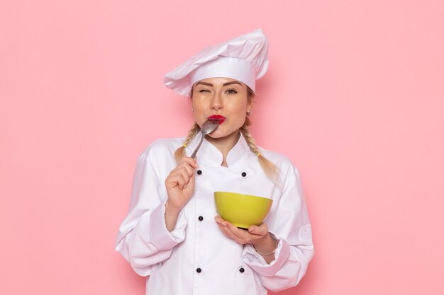 Вид спереди молодая женщина-повар в белом костюме повара позирует, улыбаясь и держа зеленую тарелку, дегустируя ее на розовом пространстве, повар кухня работа работа фото