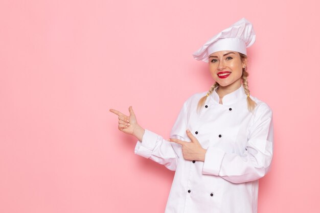 Front view young female cook in white cook suit posing and smiling on the pink space cook cuisine job work s photo