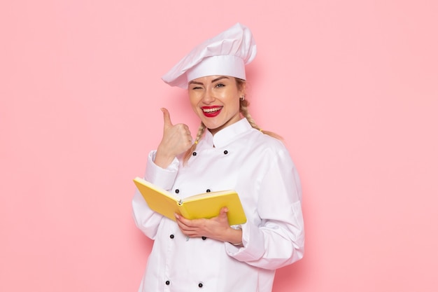 Foto gratuita giovane cuoco femminile di vista frontale in quaderno bianco della lettura del vestito del cuoco con il sorriso sul cuoco rosa dello spazio