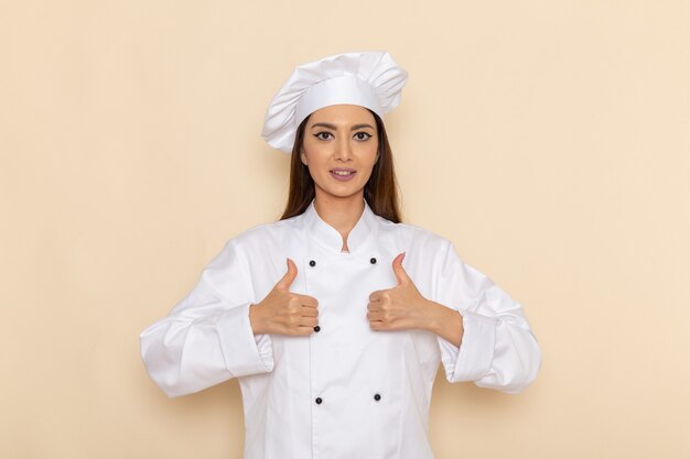 Front view of young female cook in white cook suit smiling and showing like signs on light-white wall