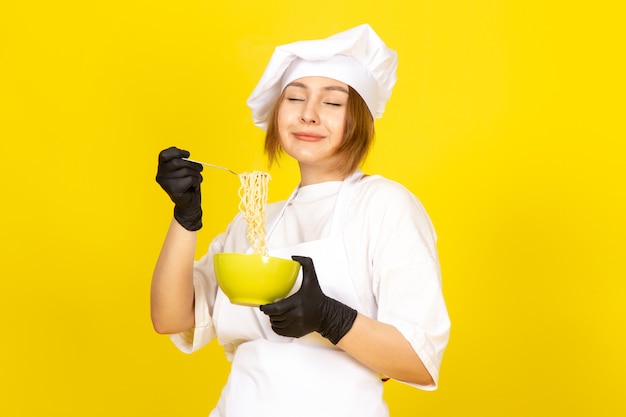白いコックスーツと黄色のスパゲッティを混合する緑のプレートを保持している黒い手袋の白い帽子の正面の若い女性料理人