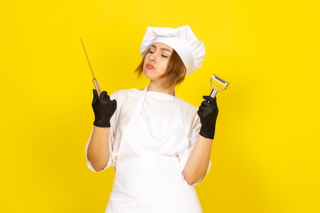 A front view young female cook in white cook suit and white cap in black gloves holding knife and vegetable cleaner on the yellow