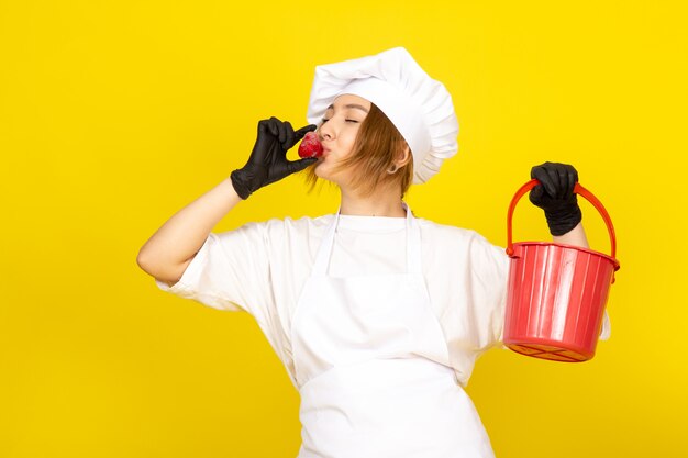 A front view young female cook in white cook suit and white cap in black gloves holding red basket and strawberry kissing it on the yellow