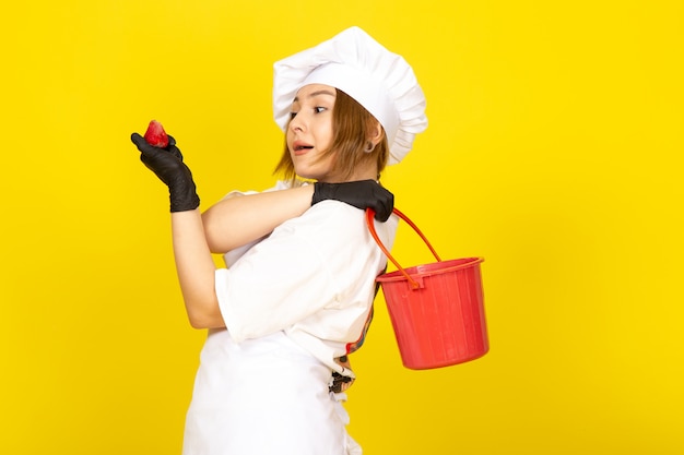 A front view young female cook in white cook suit and white cap in black gloves holding red basket and strawberry on the yellow