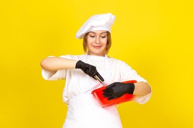 A front view young female cook in white cook suit and white cap in black gloves holding red bowl mixing it up on the yellow