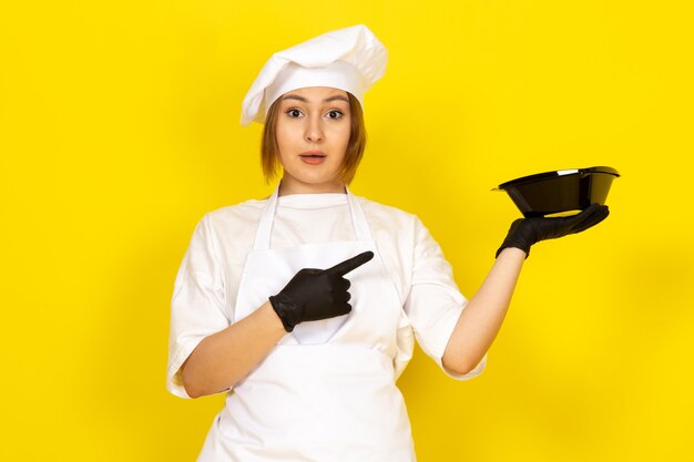 A front view young female cook in white cook suit and white cap in black gloves showing black bowl on the yellow