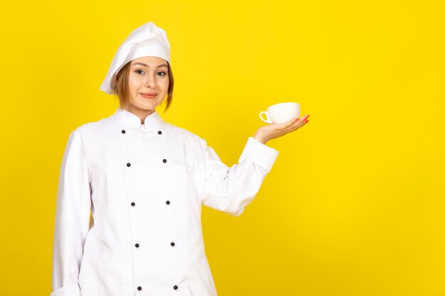 Вид спереди молодая женщина-повар в белом поварском костюме и белой кепке пьет, держа чашку кофе, улыбаясь на желтом