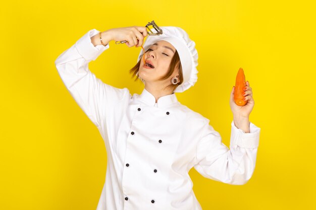 Вид спереди молодая самка повар в белом костюме повара и белой кепке держит и чистит едят оранжевую морковь на желтом