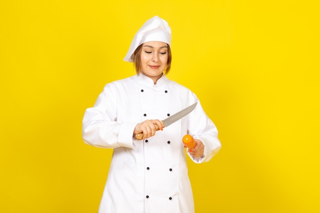 白いクックスーツと黄色のカッティングキャロットを笑顔でナイフを保持している白い帽子の正面の若い女性クック