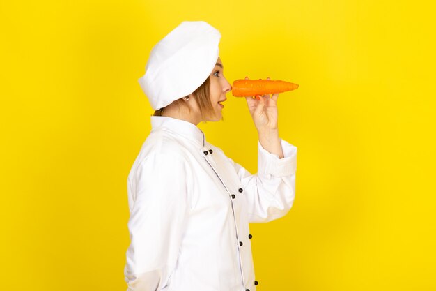 A front view young female cook in white cook suit and white cap holding orange carrot smiling funny on the yellow