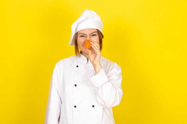 A front view young female cook in white cook suit and white cap holding orange carrot smiling funny on the yellow