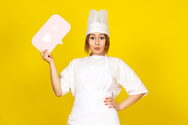 Вид спереди молодая женщина повар в белом поварском костюме и белой кепке с розовым знаком позирует на желтом