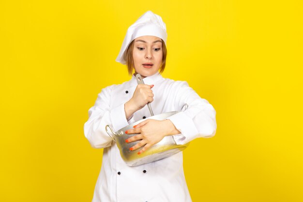 A front view young female cook in white cook suit and white cap holding round silver pan mixing it up surprised on the yellow