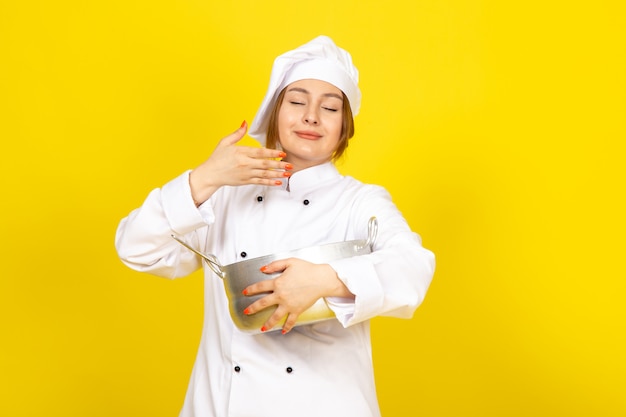 Вид спереди молодая женщина-повар в белом кухонном костюме и белой кепке, держащая круглую серебряную сковороду, пахнущую на желтом