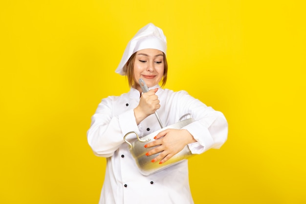 A front view young female cook in white cook suit and white cap holding round silver pan smiling on the yellow