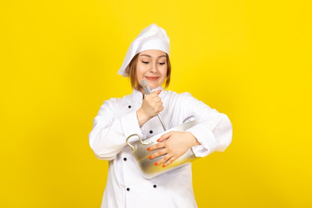 Вид спереди молодая женщина-повар в белом кухонном костюме и белой кепке, держащая круглую серебряную кастрюлю, улыбаясь на желтом