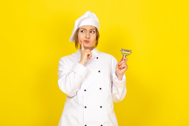 A front view young female cook in white cook suit and white cap holding vegetable cleaner posing thinking on the yellow