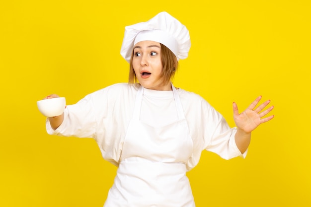 A front view young female cook in white cook suit and white cap holding white cup excited funny on the yellow