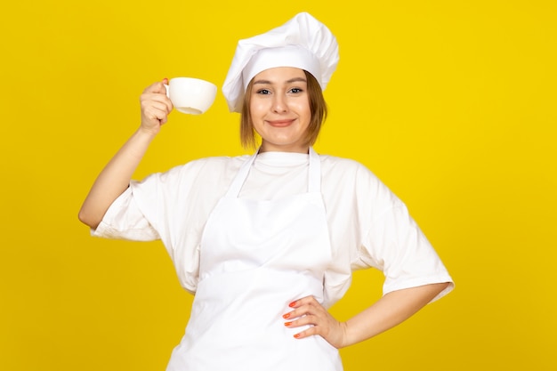 A front view young female cook in white cook suit and white cap holding white cup smiling on the yellow