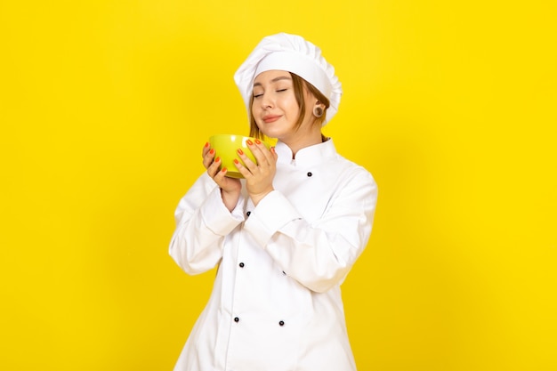 A front view young female cook in white cook suit and white cap holding yellow plate delighted on the yellow