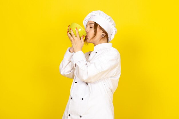 A front view young female cook in white cook suit and white cap holding yellow plate tasting on the yellow