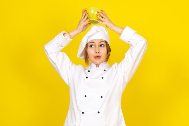A front view young female cook in white cook suit and white cap holding yellow plate on the yellow