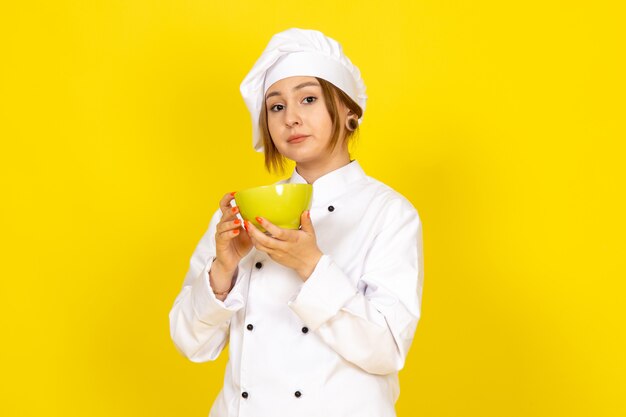 A front view young female cook in white cook suit and white cap holding yellow plate on the yellow