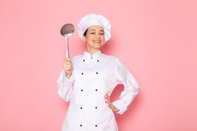 A front view young female cook in white cook suit white cap posing holding big silver spoon smiling happy expression