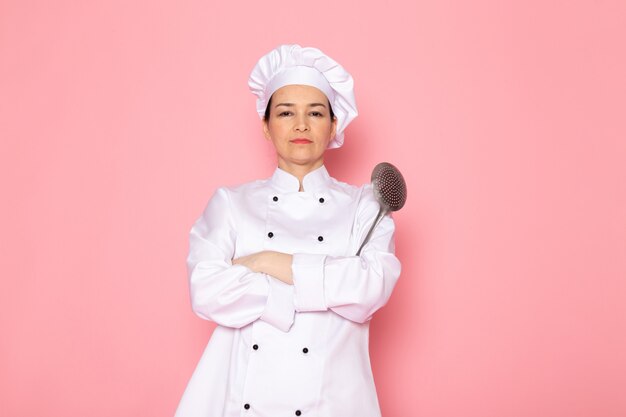 A front view young female cook in white cook suit white cap posing holding big silver spoon strict expression