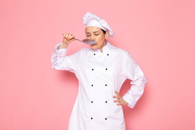 A front view young female cook in white cook suit white cap posing holding big silver spoon tastying meal