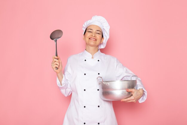 A front view young female cook in white cook suit white cap posing holding silver saucepan and big silver spoon smiling delighted