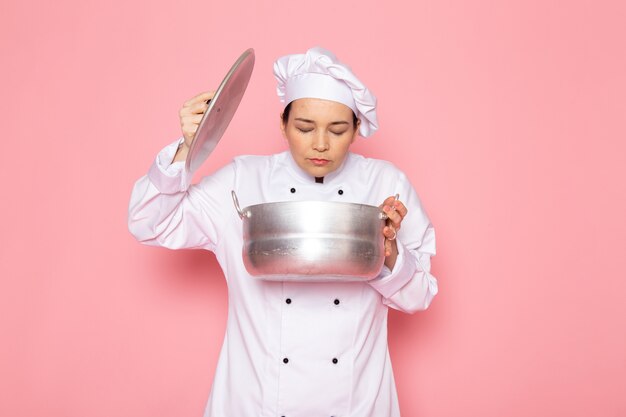 A front view young female cook in white cook suit white cap posing holding silver saucepan smelling inside