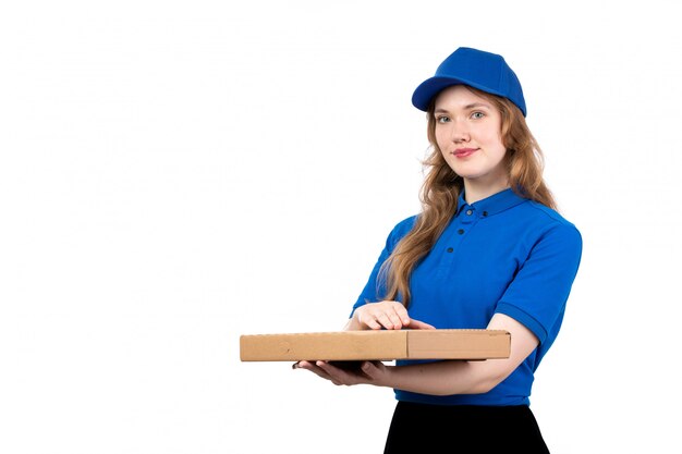 A front view young female courier in blue shirt blue cap and black trousers holding delivery box smiling on white