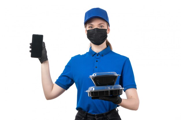 A front view young female courier in blue uniform black gloves and black mask holding food delivery bowls and phone